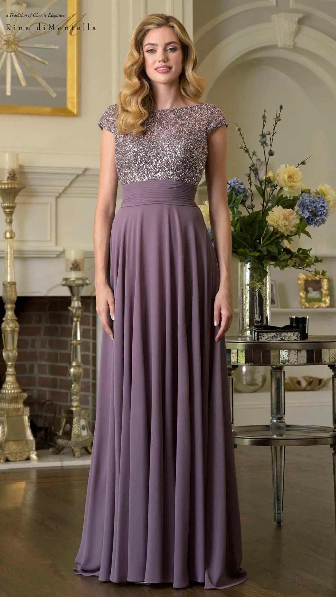 A woman indoors wears the Rina Di Montella RD3419 long formal dress in Victorian Lilac, featuring a beaded bodice and glittery fitted top with cap sleeves, surrounded by elegant dรฉcor and flowers.